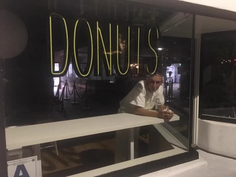 Marc at night, leaning on the counter of a walk-up window under a neon sign that reads DONUTS, in a white T-shirt, looking out at the camera.