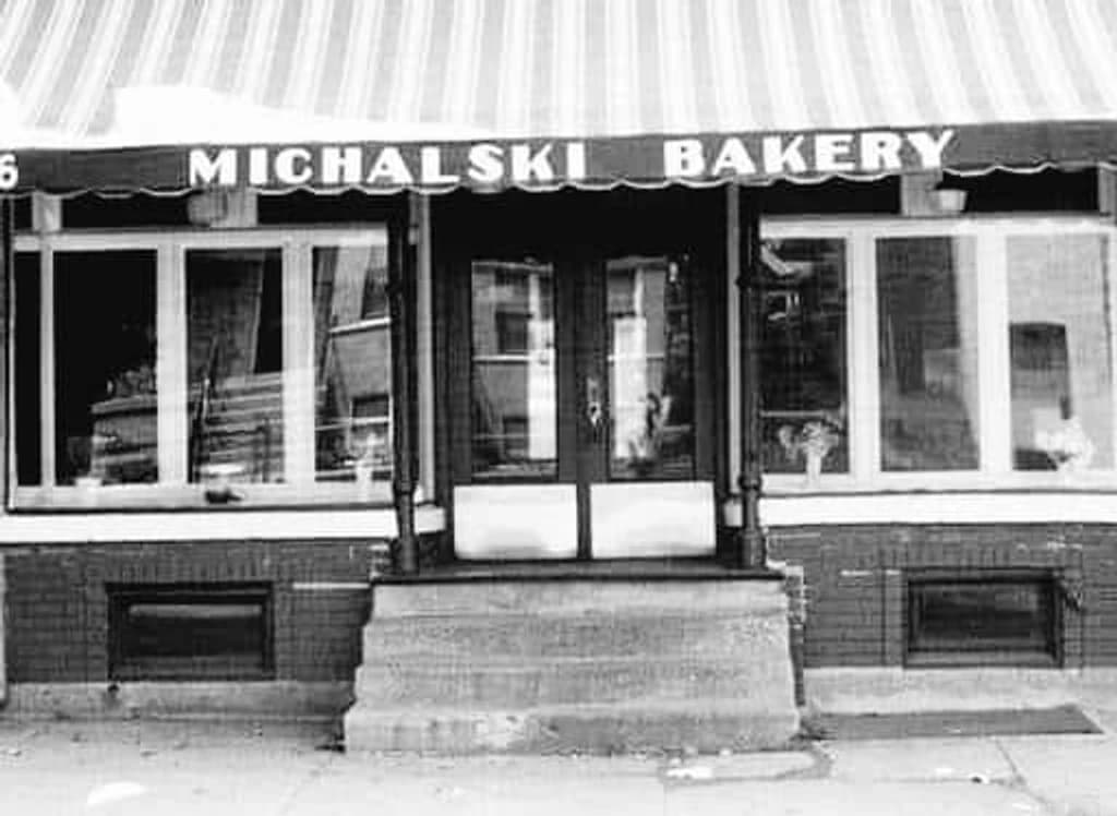 A black and white photograph of the Michalski Bakery storefront: a striped awning over three plate-glass windows, MICHALSKI BAKERY lettered across the front, and a double door at the top of a short flight of stone steps.